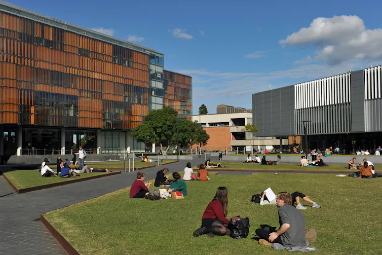 People sit on a lawn, surrounded by buildings.