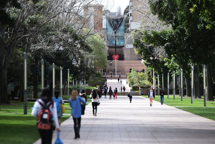 People walk on a broad walkway, surrounded by trees. A multi-story building is in the background.