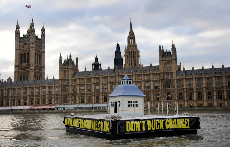 A barge floating past the houses of parliament with a sign reading 'don't duck change' on the side.