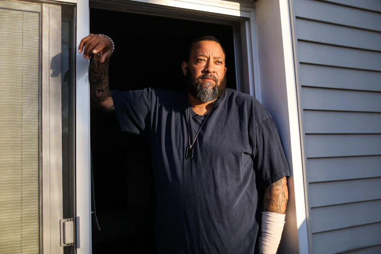 A Black man stands in a doorway with a heavily bandaged arm and stares into the distance.