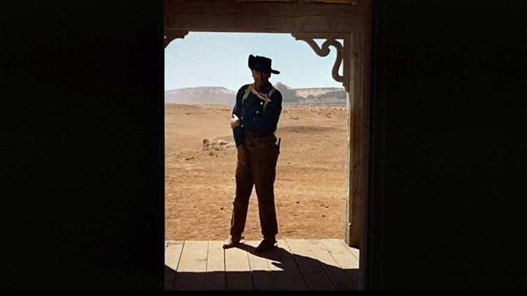 John Wayne stands in a doorway, with a desert stretching into the distance behind him.