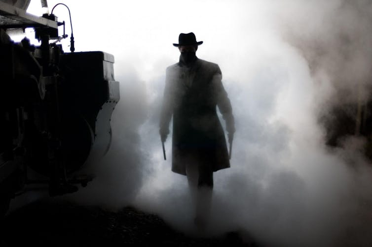 A man wearing a long coat, hat and face mask, and carrying two pistols, emerges from a cloud of smoke.