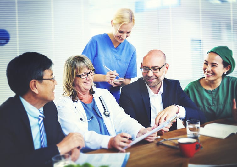 Five people collaborating in a meeting room, two in business attire and three in clinical clothes
