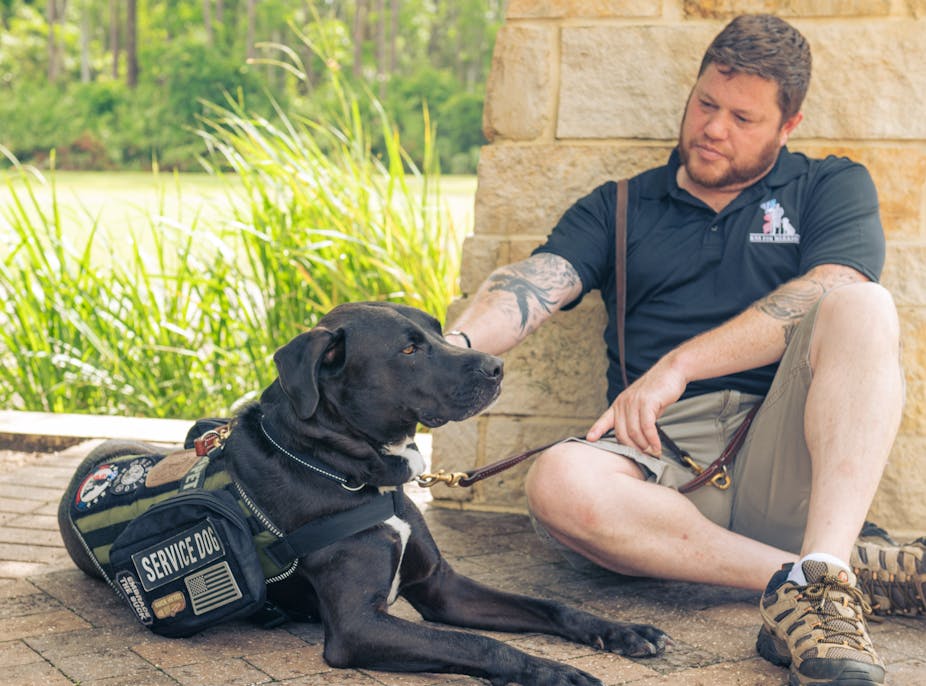 Man sits on the ground with a service dog wearing a vest.