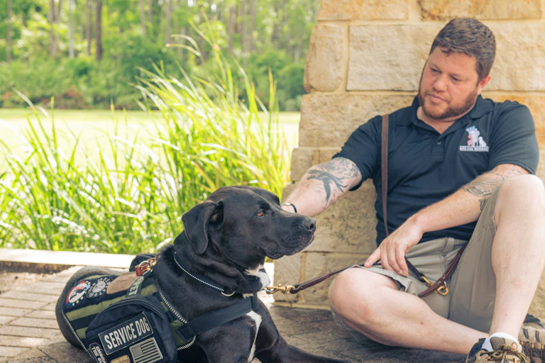 Man sits on the ground with a service dog wearing a vest.