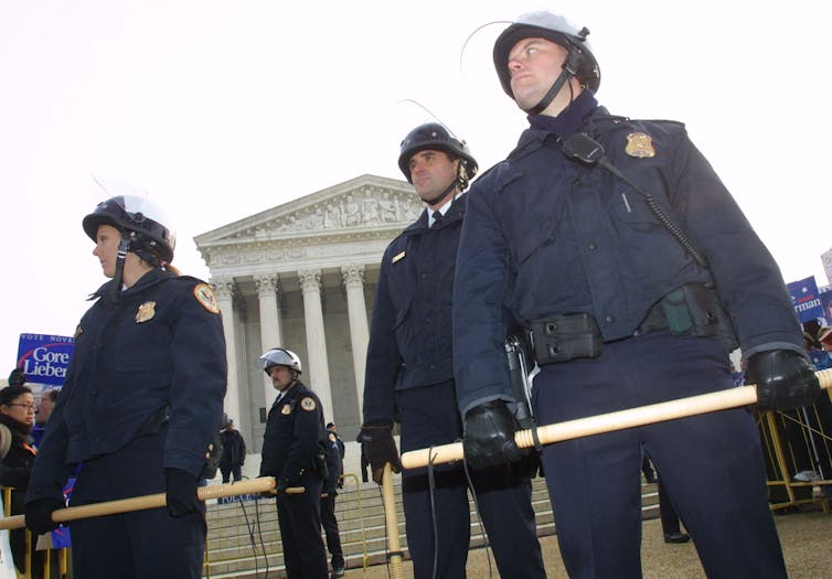 Police officers in riot gear outside a large, white building with pillars.