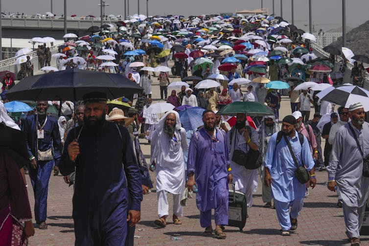 A large number of people in traditional clothing covering them from their necks to their wrists and ankles walk on wide pathway, some carrying umbrellas for shade.