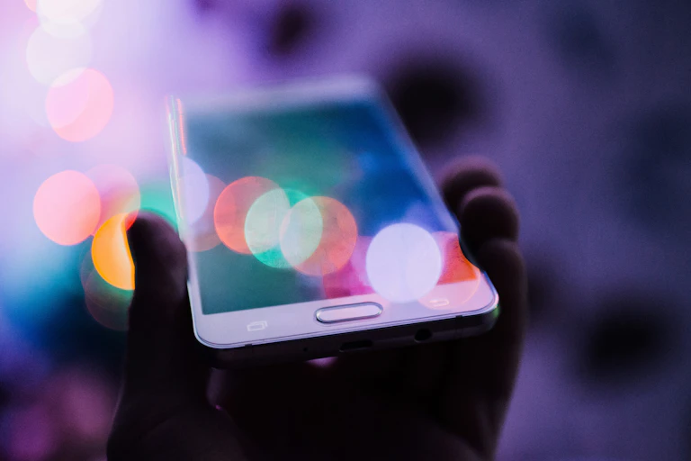 A hand holds a white smart phone under blurred coloured lights, against a shadowy, purple background.