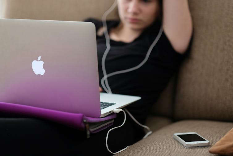 A young person sits on a couch. They have a laptop on their lap and earphones with long cords in their ears. A mobile phone lies beside them.