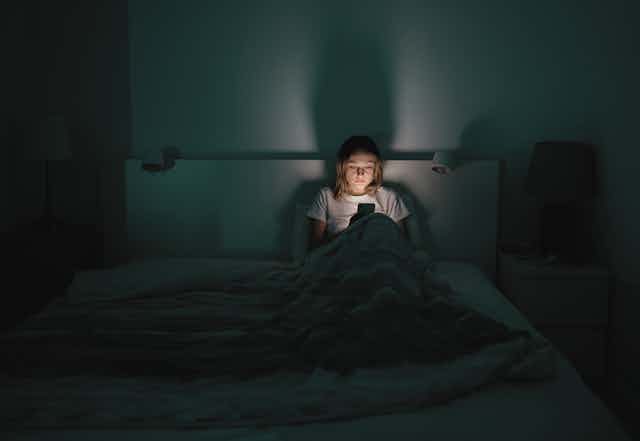 A medium distance view of a tired woman sitting up at night in her bed looking at a smartphone.