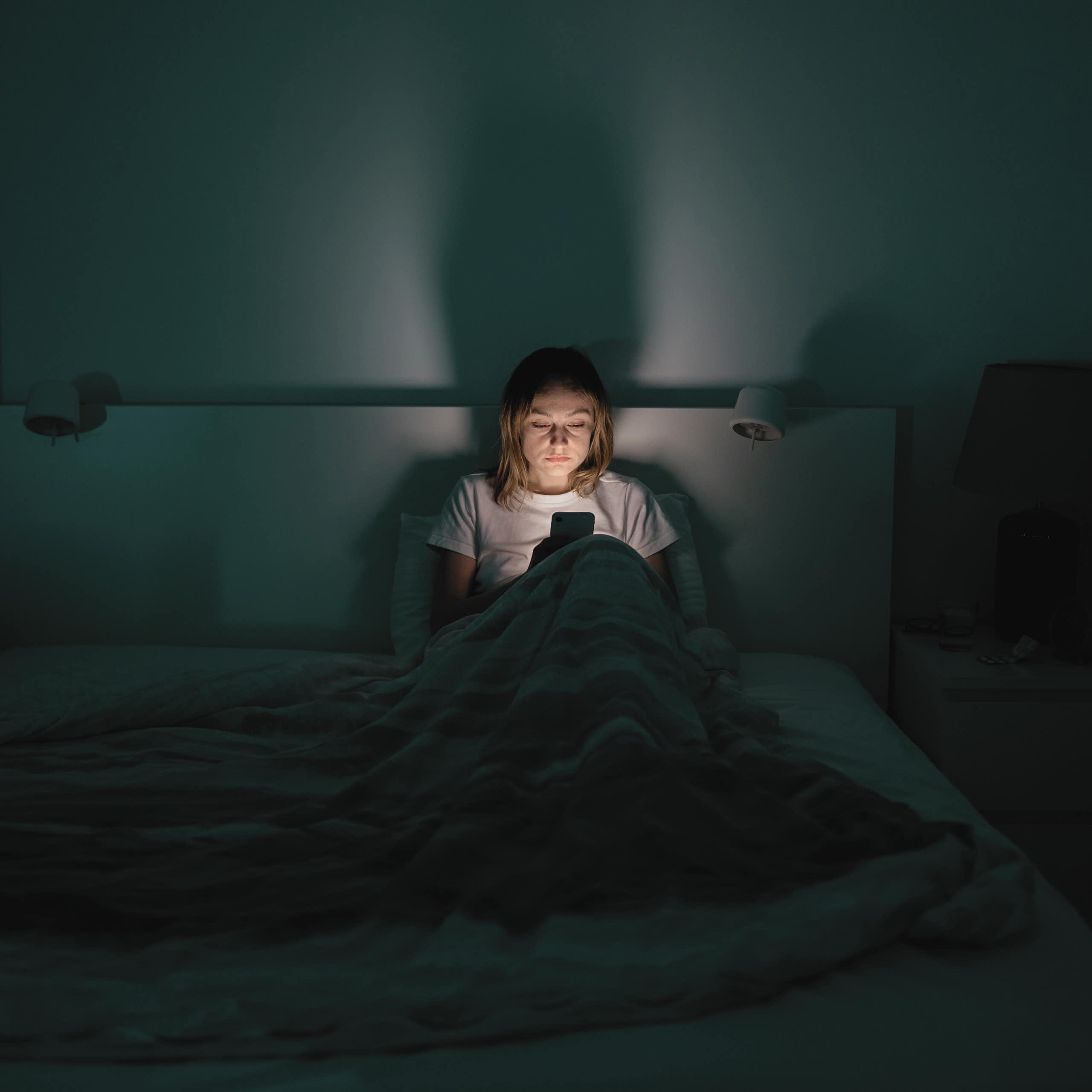 A medium distance view of a tired woman sitting up at night in her bed looking at a smartphone.