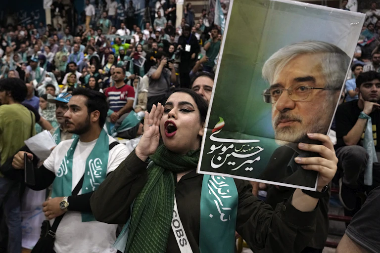 A large crowd of Iranian supporters of Masoud Pezeshkian gather in Tehran; in the foreground, a young woman shouts her endorsement while holding up a campaign sign.