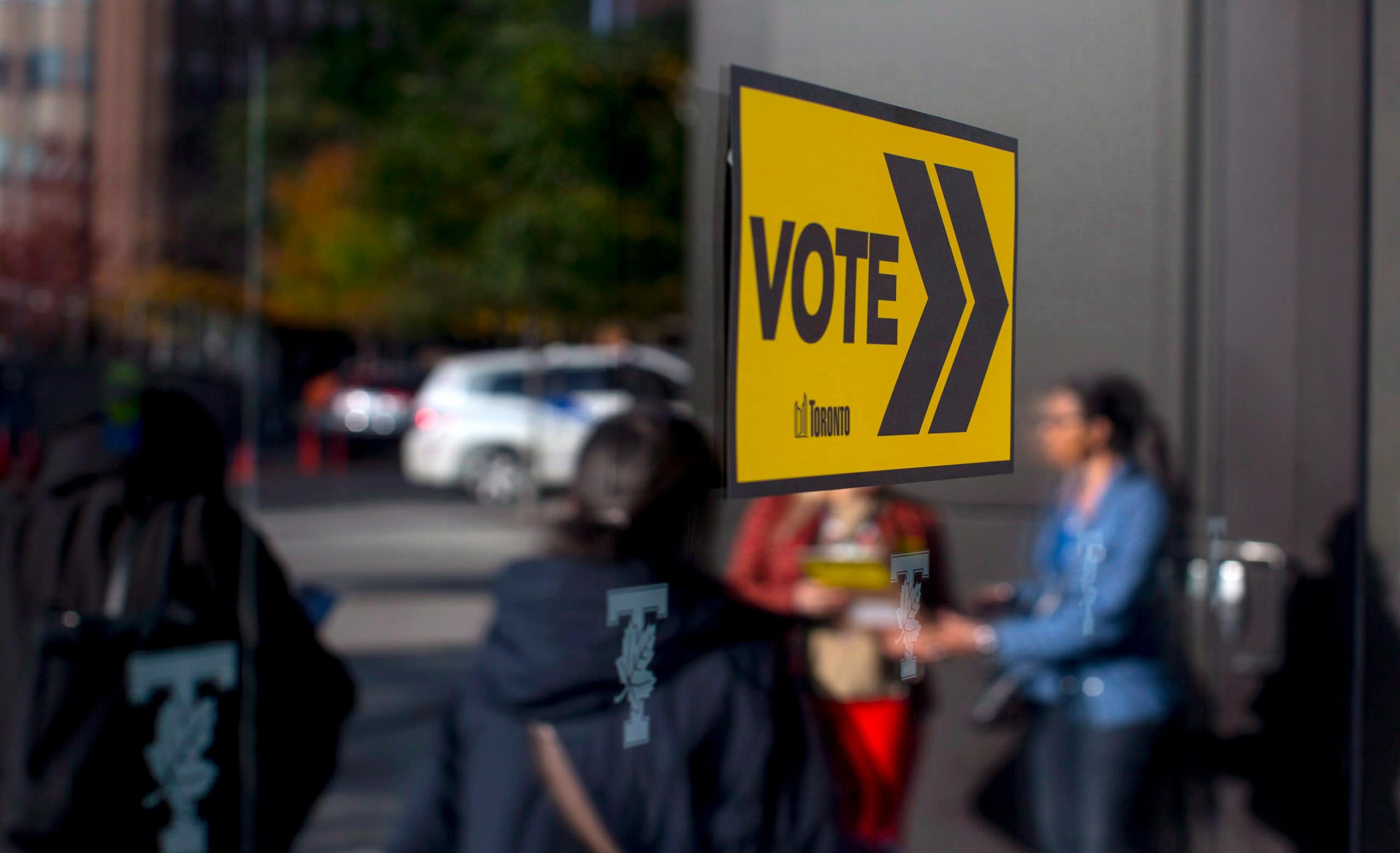 A yellow Vote sign is attached to glass; voters are reflected in the glass.