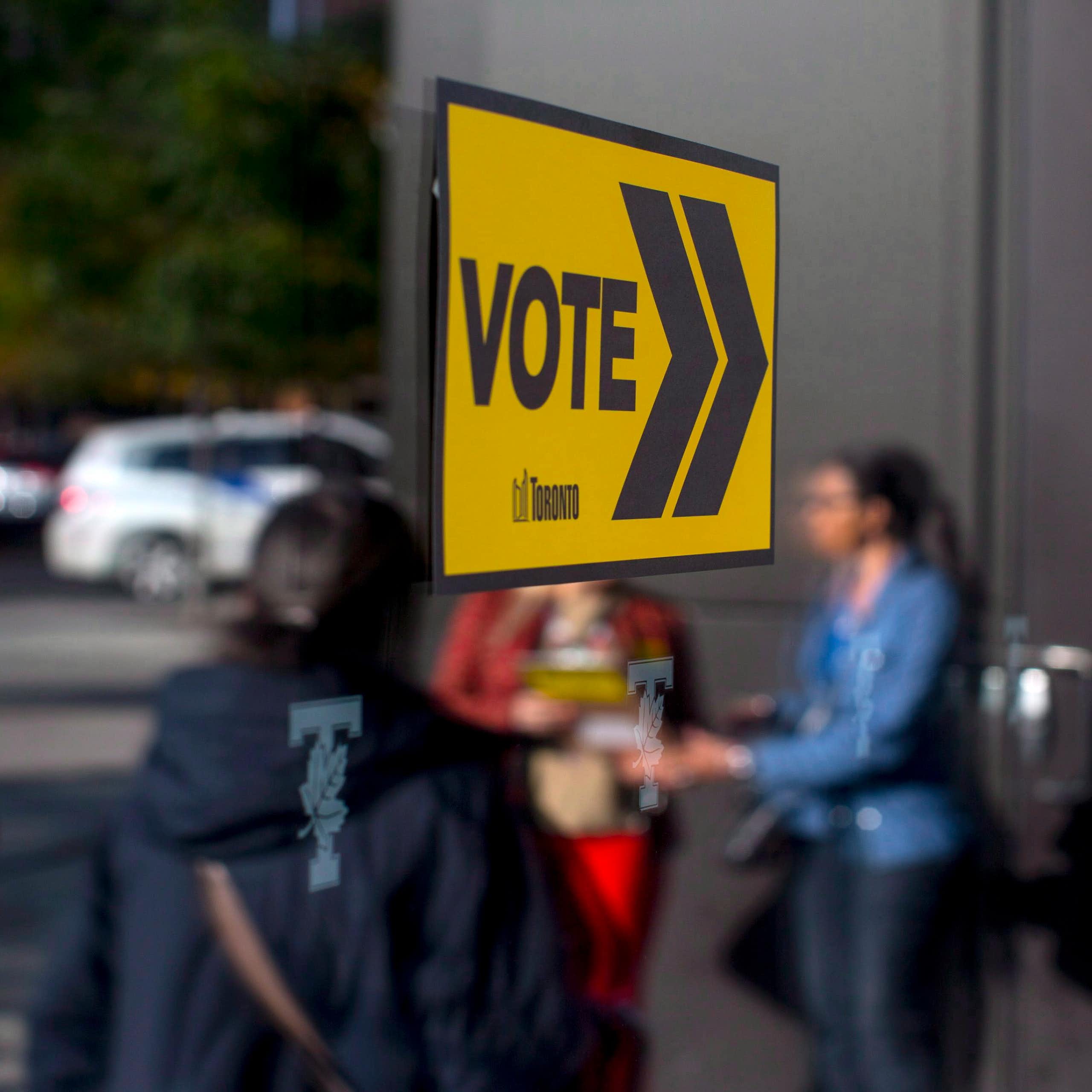 A yellow Vote sign is attached to glass; voters are reflected in the glass.