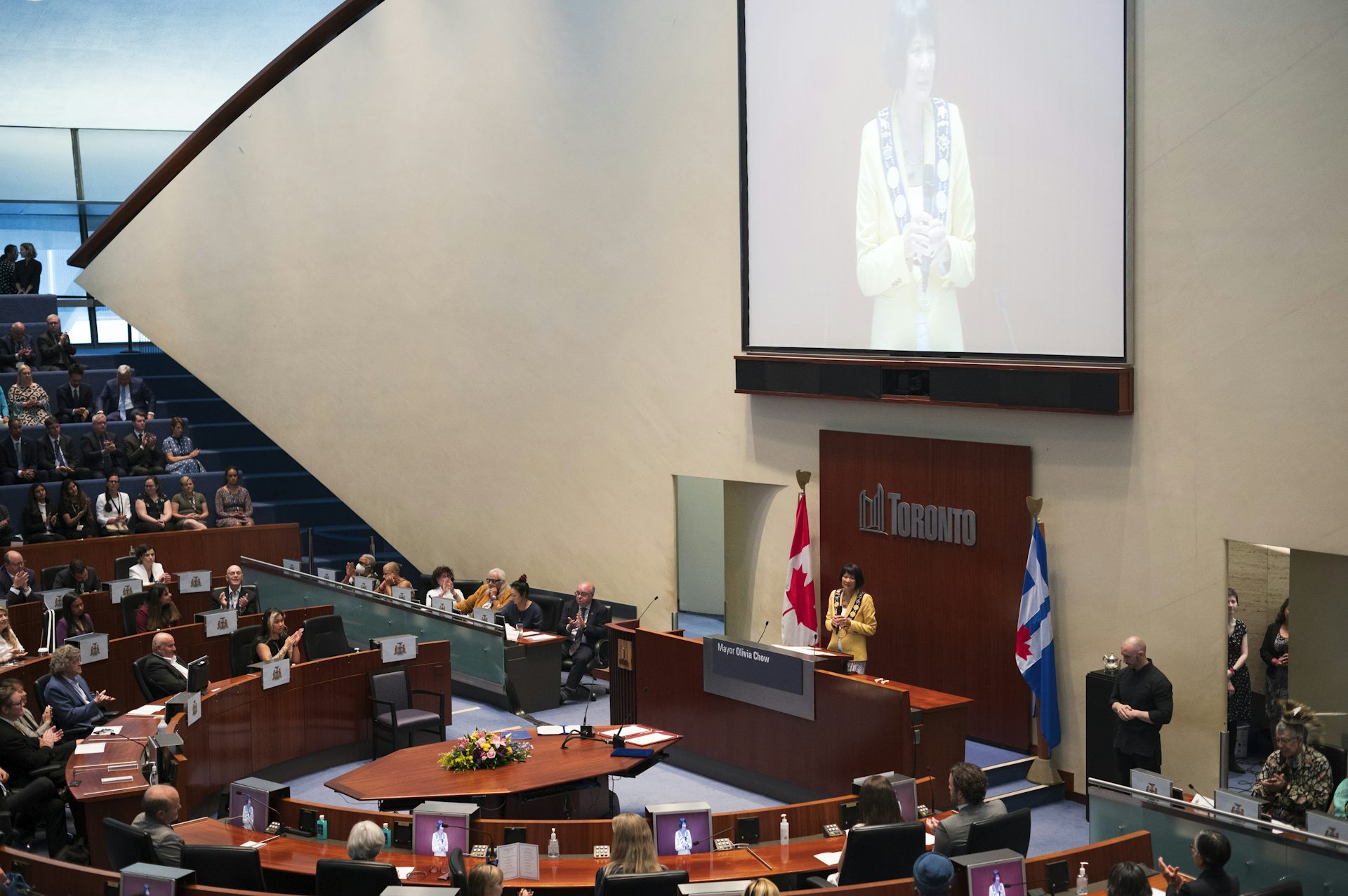 A woman beneath a big screen of her image in city council chambers.