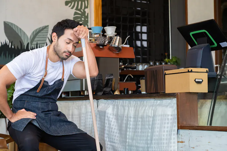Man wearing an apron, sitting down resting his forehead on the end of a broom, looking tired.