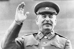 A black and white photo shows a smiling man with a moustache in a military uniform raising his right hand.