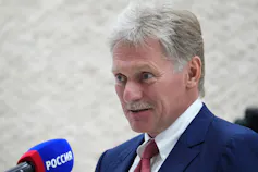 A grey-haired man in a blue jacket smiles faintly while speaking into a microphone.