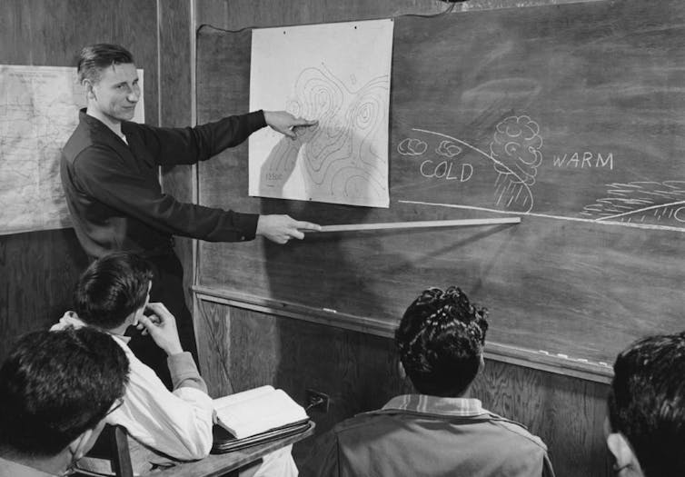 Black and white photo of a young teacher pointing at a blackboard while students watch from their desks.
