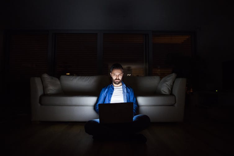 A man is sitting on the floor in a dark room, leaning on a sofa and using a laptop.