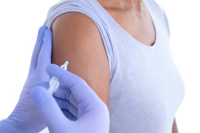 Une femme reçoit une injection dans le bras par une paire de mains portant des gants violets.
