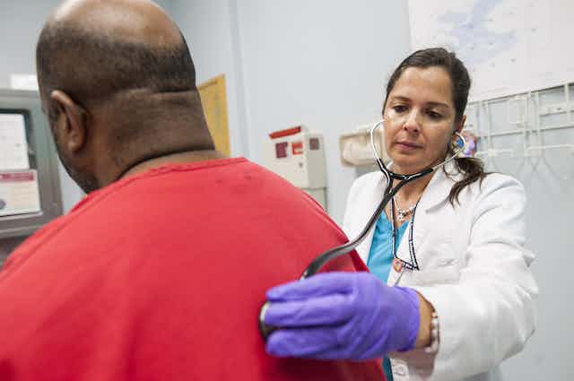 A woman in a white coat presses a stethoscope to the back of a man wearing a red shirt