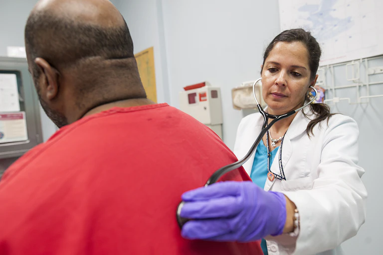 A woman in a white coat presses a stethoscope to the back of a man wearing a red shirt
