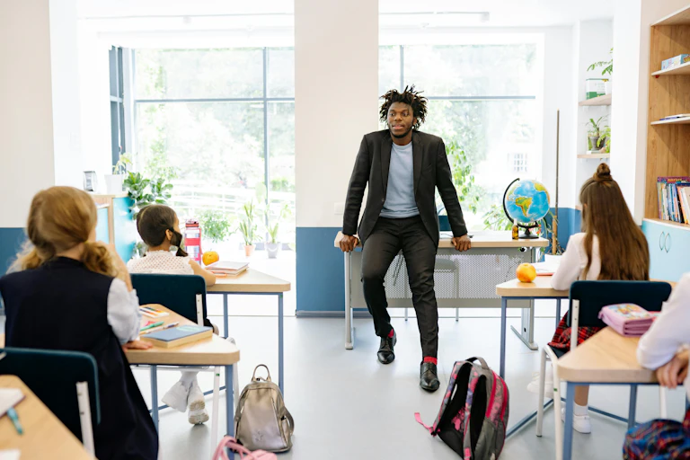 A man sits on a desk at the front of room, speaking to children un uniforms, seated at tables in front of him.
