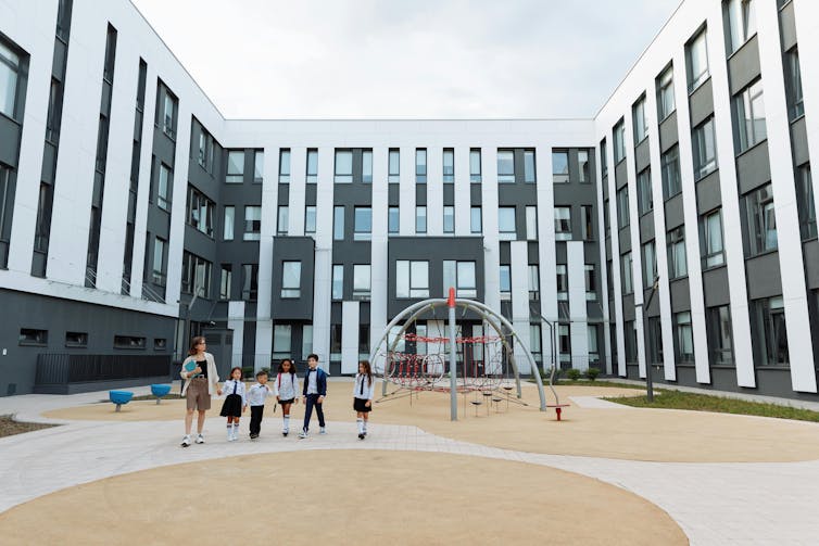 A woman walks a group of children in uniform past play equipment. They are in a courtyard surrounded by a modern four-story building.