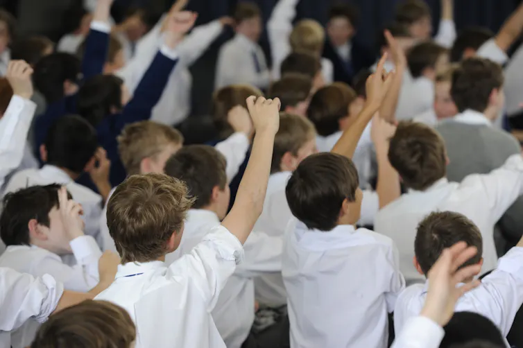 Boys in white shirts sit on the floor. Some have their hands raised.