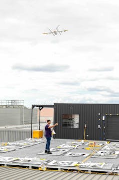 Photo of a man standing on a launchpad full of drones, attaching a box to a cord leading up