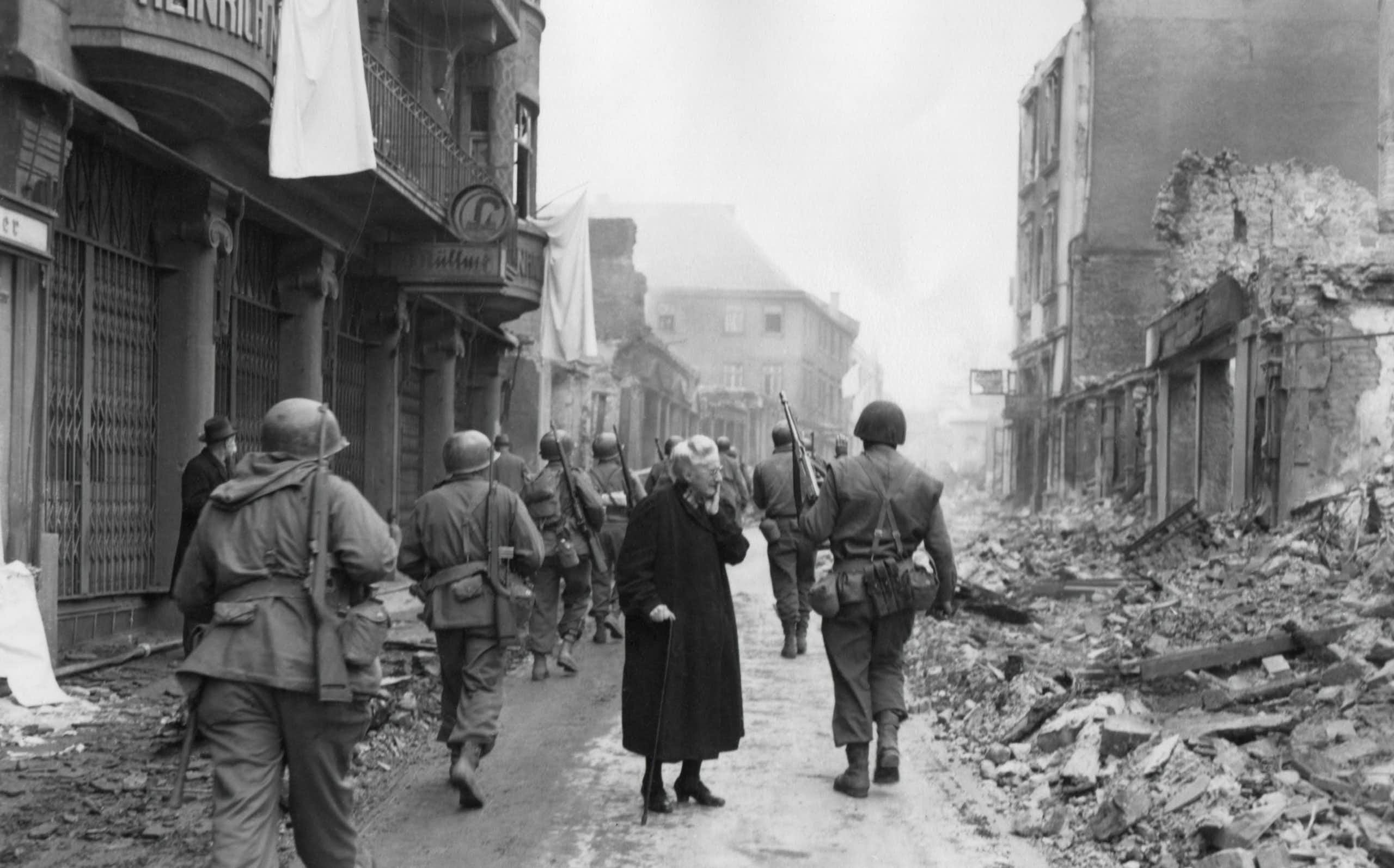 As infantrymen march through a German town, a shocked old woman stares at a the ruins.