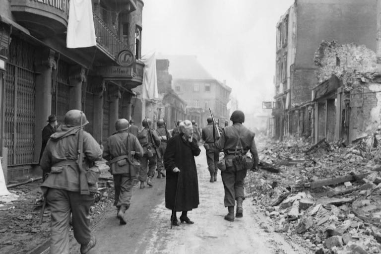 As infantrymen march through a German town, a shocked old woman stares at a the ruins.