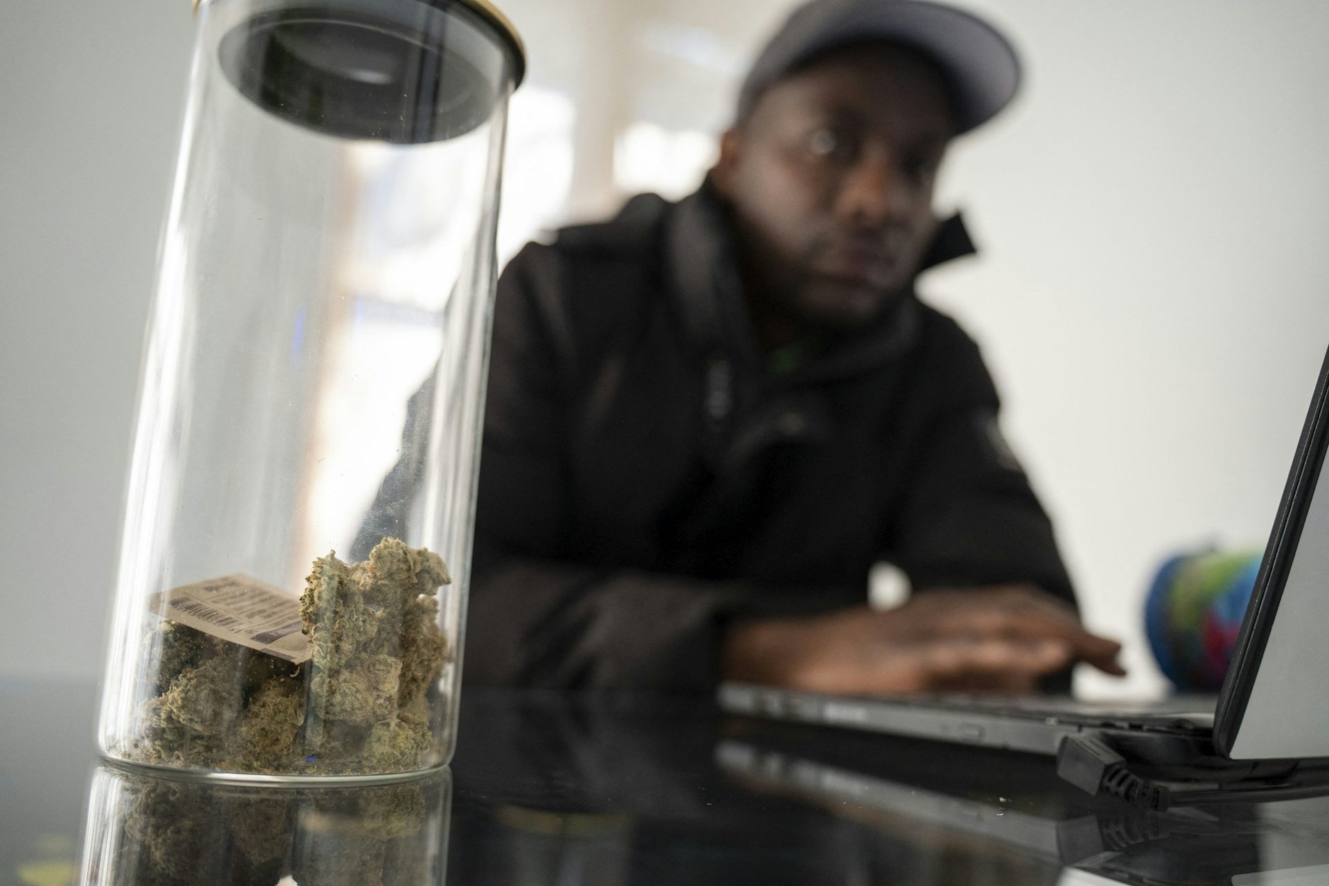 Au premier plan, un flacon contient de la résine de cannabis. En arrière-plan, on voit un jeune homme africain avec une casquette sur la tête