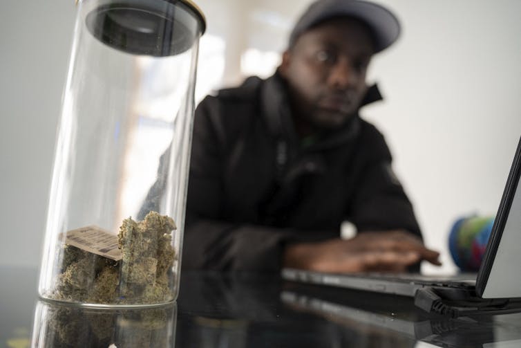Au premier plan, un flacon contient de la résine de cannabis. En arrière-plan, on voit un jeune homme africain avec une casquette sur la tête