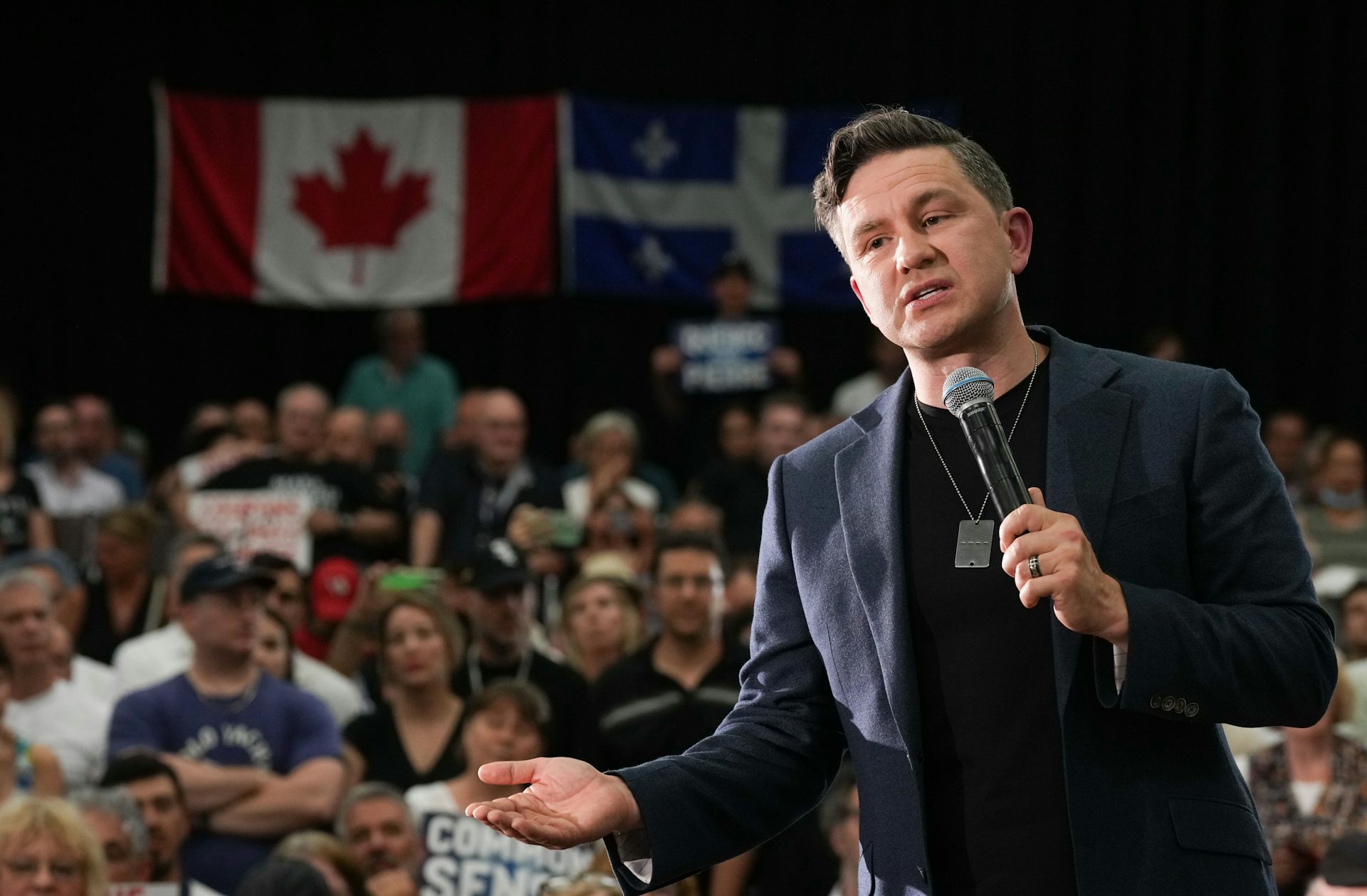 A dark-haired man holds a microphone at a rally, speaking to a crowd as a large Canadian flag hangs behind him.