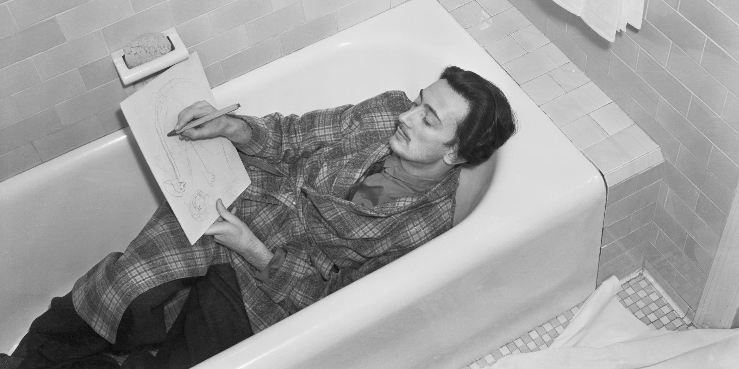 Black and white photo of young man with thin mustache lying in a bathtub, fully clothed, while sketching on a piece of paper.