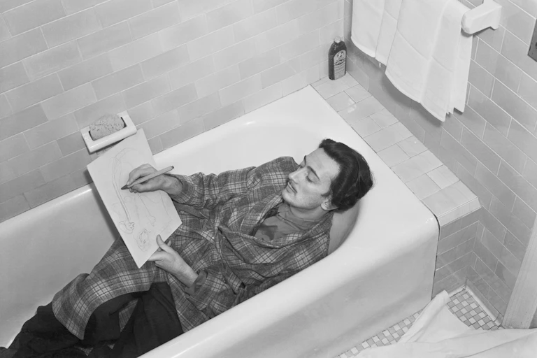Black and white photo of young man with thin mustache lying in a bathtub, fully clothed, while sketching on a piece of paper.