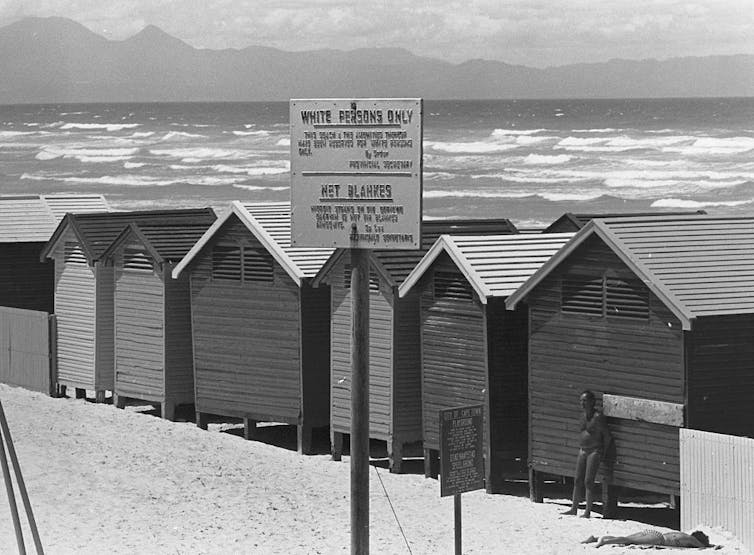 A black and white photo of old beach huts, a man leaning against one. A sign reads