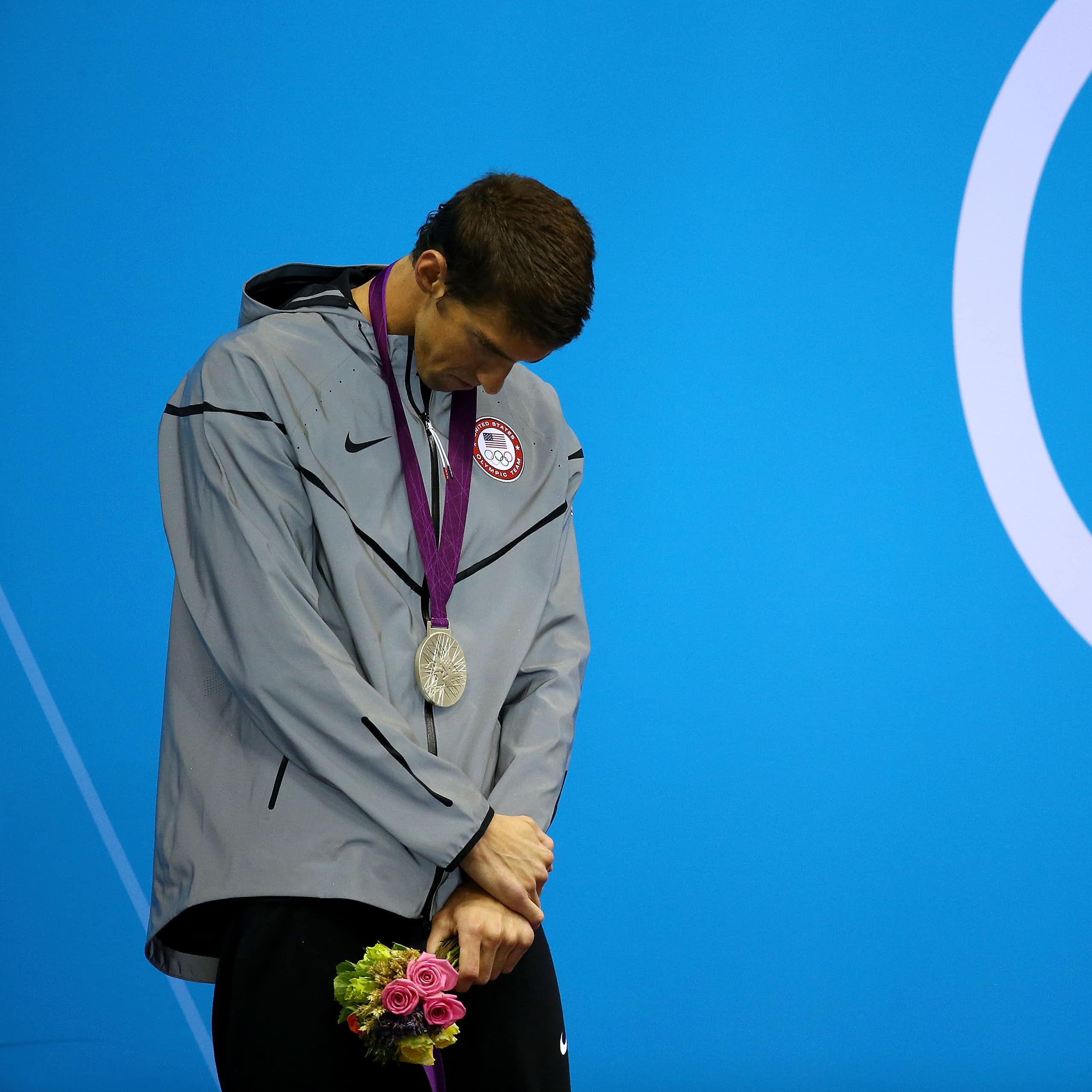 Young man hanging his head while wearing a gray windbreaker with a silver medal around his neck.