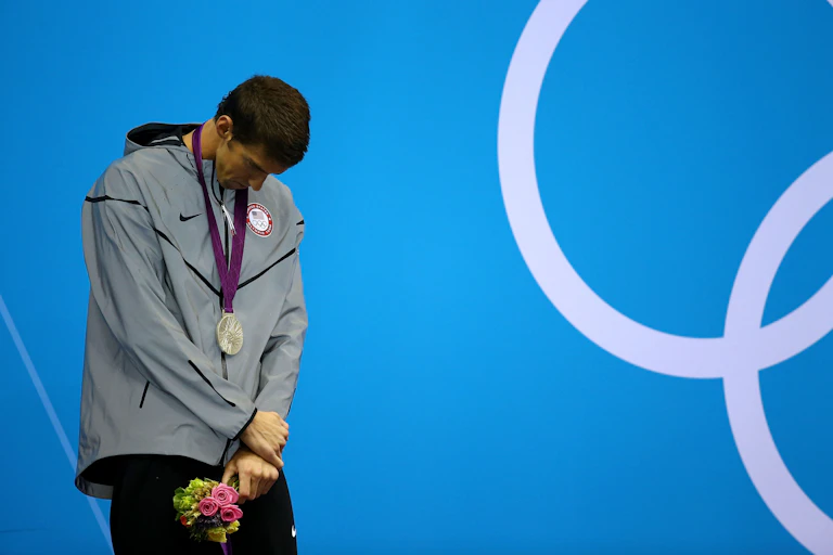 Young man hanging his head while wearing a gray windbreaker with a silver medal around his neck.