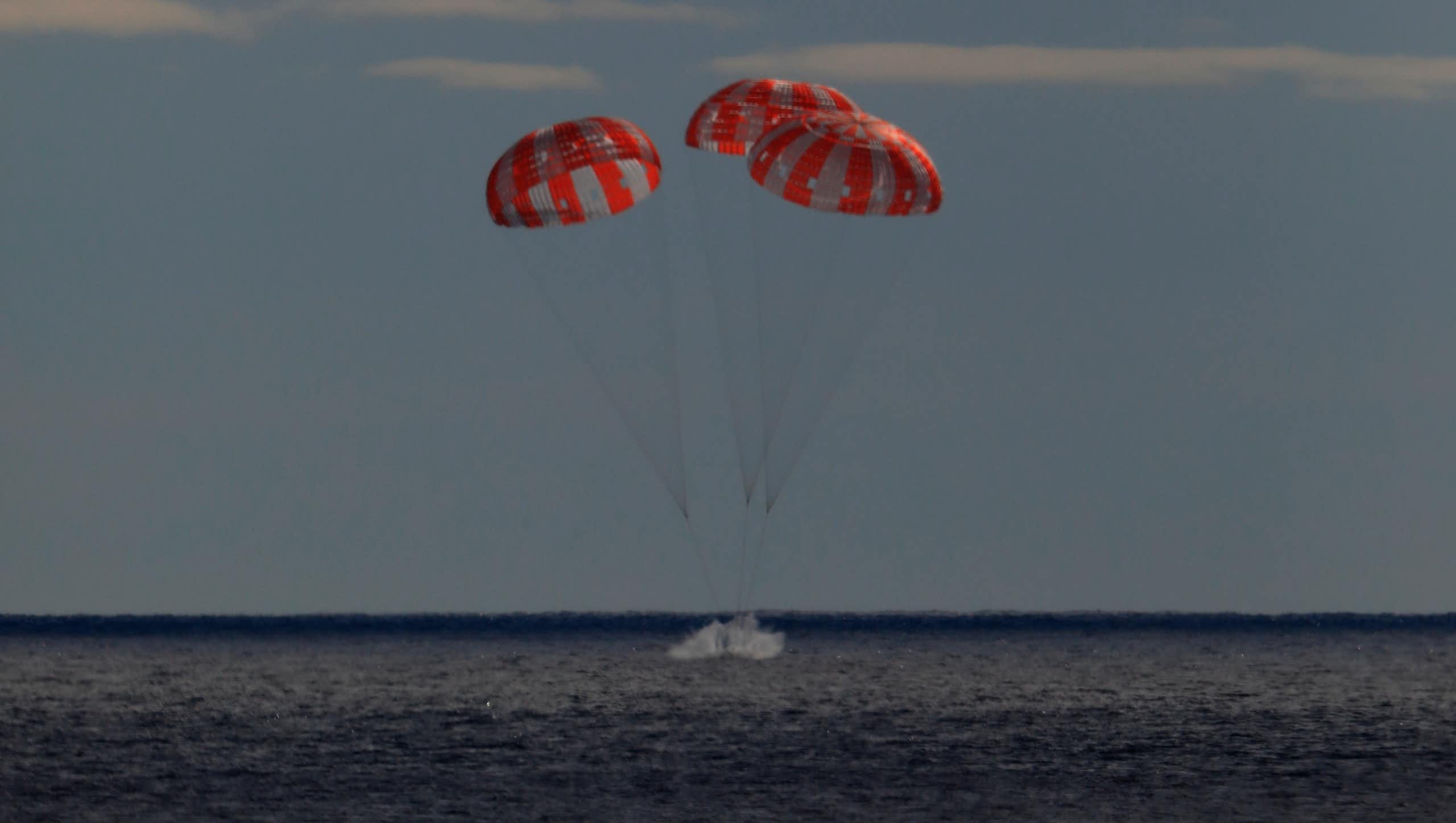 A small metal capsule carried by three parachutes lands in a body of water. 