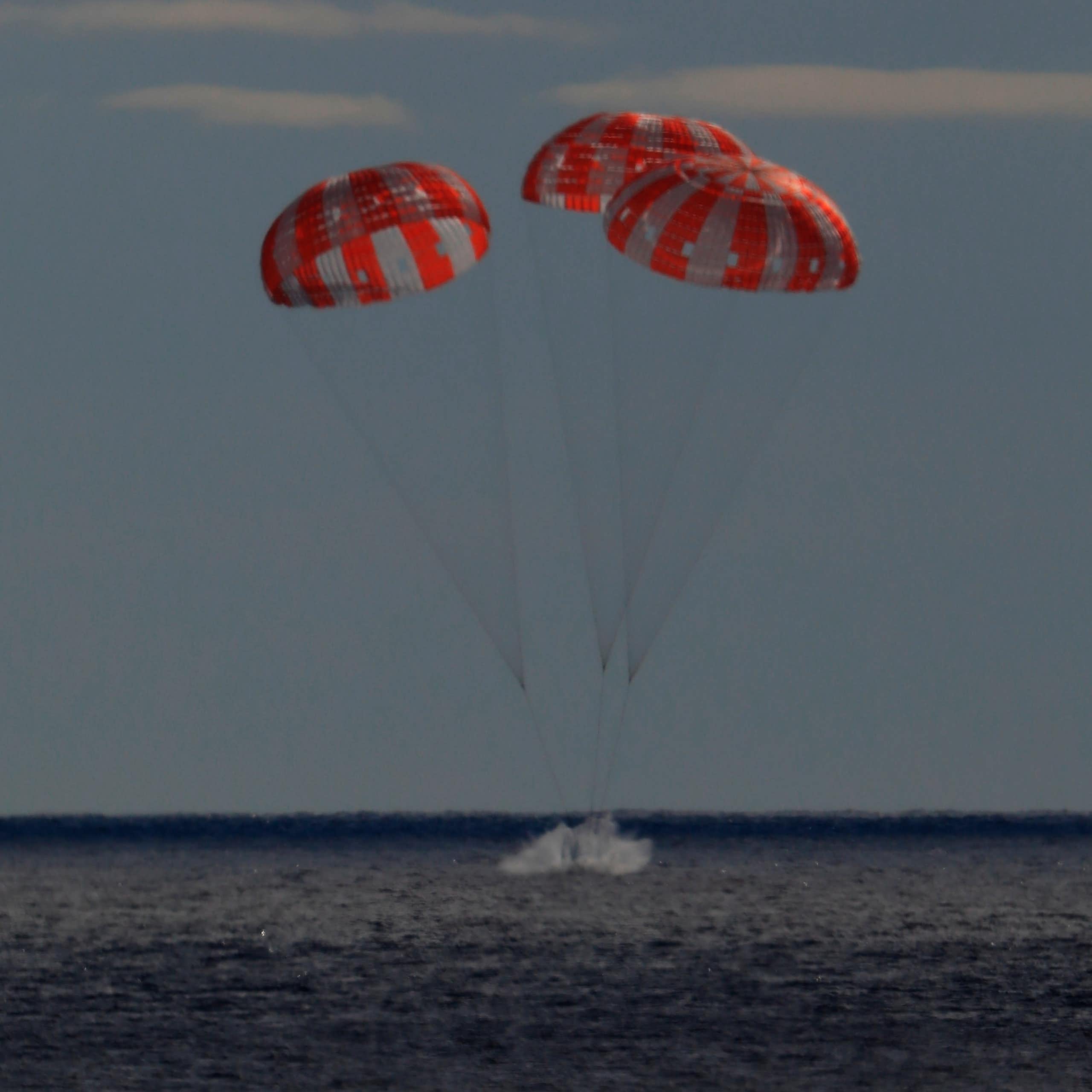 A small metal capsule carried by three parachutes lands in a body of water. 