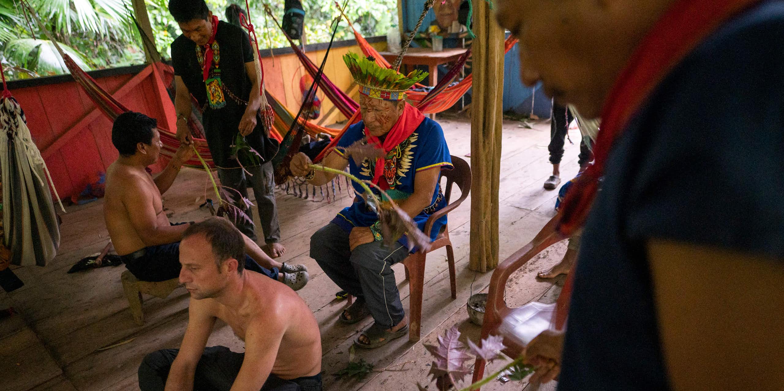 Two bare-chested men sit on the floor, while two others wearing traditional dress wave a plant near them.