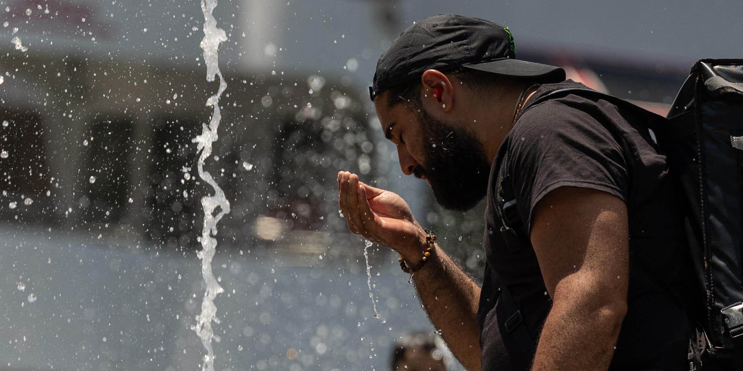 A man cools his face with water from a fountain while a boy splashes in the water behind him.