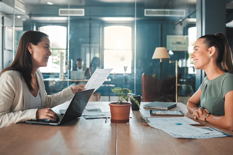two businesswomen sit across a desk from each other and discuss documents