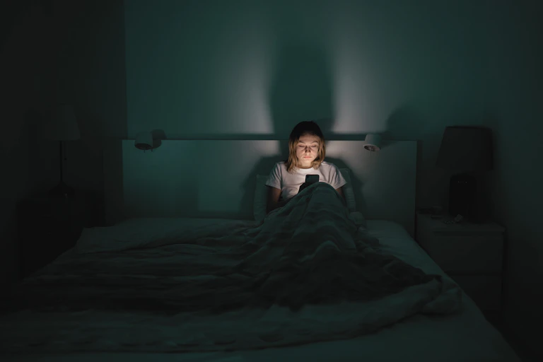Uma visão de média distância de uma mulher cansada sentada à noite em sua cama, olhando para um smartphone