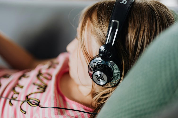 An adolescent lies with headphones on. Their heads is propped up by a cushion.