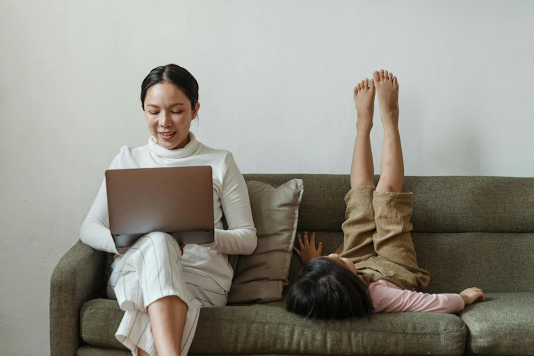 A woman sits on a couch with a laptop. A child lies next to the woman, with her feet in the air.