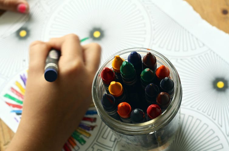 A child colours in a sheet. A jar of crayons sits near their hand.